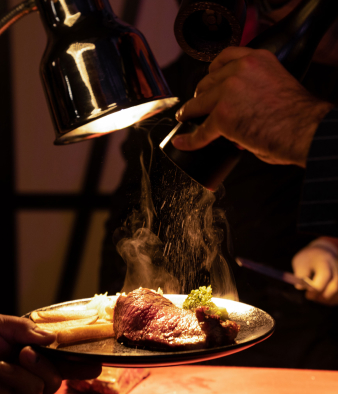 Steak auf einem Teller unter einer Warmhalte-Lampe welches gewürzt wird